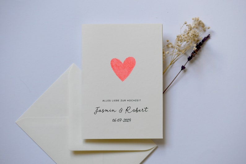 May include: A white card with a pink heart and the text "Alles Liebe zur Hochzeit"  "Jasmin & Robert" "06.07.2024".