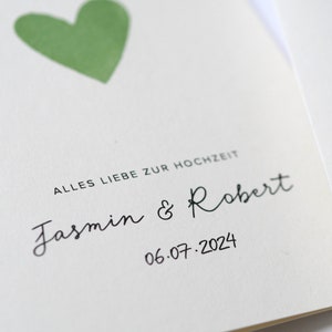 May include: A white card with a green heart and the text "Alles Liebe zur Hochzeit" in black. Below the heart, the names "Jasmin & Robert" are written in cursive, followed by the date "06.07.2024".