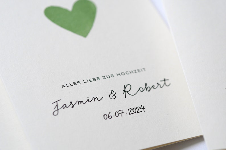 May include: A white card with a green heart and the text "Alles Liebe zur Hochzeit" in black. Below the heart, the names "Jasmin & Robert" are written in cursive, followed by the date "06.07.2024".