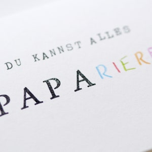 以下が含まれることがあります： 白いカードに「Du kannst alles  PAPARIEREN」という黒い文字とカラフルな文字が書かれています。 「PAPARIEREN」という単語は、それぞれの文字が異なる色で書かれています。