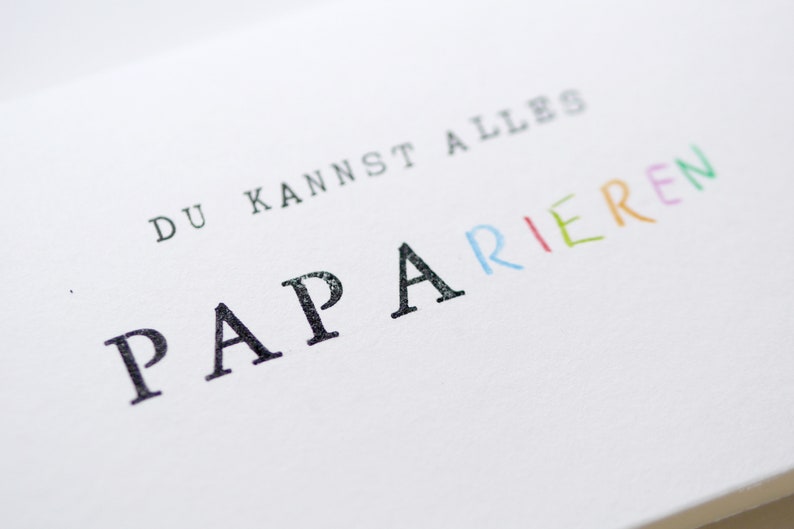 以下が含まれることがあります： 白いカードに「Du kannst alles  PAPARIEREN」という黒い文字とカラフルな文字が書かれています。 「PAPARIEREN」という単語は、それぞれの文字が異なる色で書かれています。