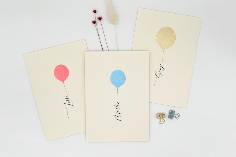 May include: Three cards with a balloon design and a name written on each. The balloons are gold, blue, and pink. The names are "Singa", "Martie", and "Jitte".