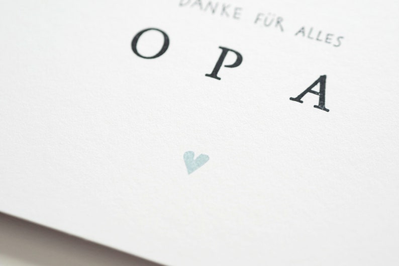 Op de afbeelding: Een witte kaart met het woord "OPA" in zwarte letters gedrukt. Het woord "Danke f&uuml;r alles" is boven het woord "OPA" gedrukt. Een klein lichtblauw hartje is onder het woord "OPA" gedrukt.