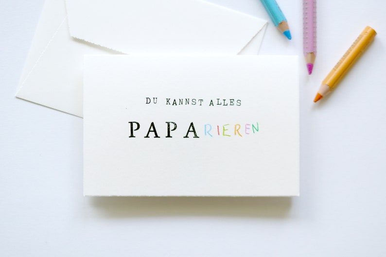 以下が含まれることがあります： 白地に黒とカラフルな文字で「Du kannst alles Papa rieren」と書かれたカード。パパという単語は虹色です。