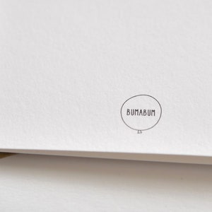 May include: A white card with a textured surface, featuring a small, circular logo with the word "BUMABUM" inside a balloon outline. The card is partially visible, with a brown envelope edge in the corner.