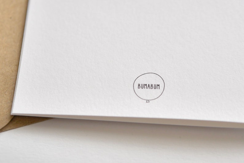May include: A white card with a textured surface, featuring a small, circular logo with the word "BUMABUM" inside a balloon outline. The card is partially visible, with a brown envelope edge in the corner.