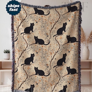 Könnte beinhalten: Eine gewebte Wohndecke mit einem sich wiederholenden Muster aus schwarzen Katzen, die auf dünnen, braunen Ästen mit kleinen orangefarbenen Blüten vor einem hellbeigen Hintergrund sitzen. Die Decke hat einen dunkelblauen Fransenrand.