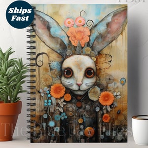 May include: A spiral bound notebook with a colorful illustration of a rabbit with large eyes and orange flowers. The rabbit is wearing a blue and brown patterned jacket. The background is a watercolor wash of yellow, orange, and blue.