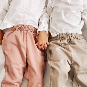 May include: Two children wearing white shirts and pants, holding hands. The child on the left is wearing pink pants, and the child on the right is wearing beige pants.