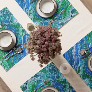 Pode incluir: Vista aérea de uma mesa com quatro individuais com um design abstrato azul e verde. Cada lugar inclui um prato branco, uma tigela, talheres e um vaso pequeno. Uma peça central de flores roxas está no meio.
