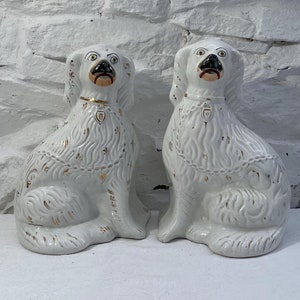 Op de afbeelding: Een paar witte keramische hondenbeeldjes met gouden accenten. De honden zitten met hun hoofd omhoog gekanteld en hun mond lichtjes open.