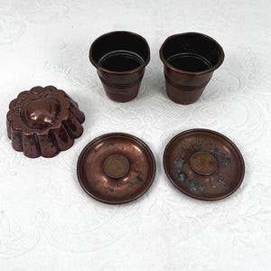 May include: An assortment of antique copper kitchenware, including two small cups, two saucers with coins, and a decorative mold. The items are arranged on a white surface, showcasing their aged patina and intricate details.