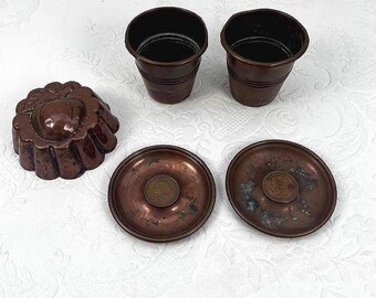 Puede incluir: Un surtido de utensilios de cocina antiguos de cobre, que incluyen dos tazas pequeñas, dos platillos con monedas y un molde decorativo. Los artículos están dispuestos sobre una superficie blanca, mostrando su pátina envejecida y detalles intrincados.