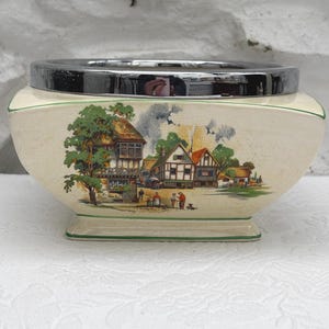 Könnte beinhalten: Cremefarbener Keramiktopf mit schwarzem Metallrand. Der Topf zeigt eine bemalte Dorfszene mit Bäumen, Häusern und Menschen. Der Boden und der Rand haben grüne Verzierungen. Der Topf steht auf einer weißen Oberfläche.