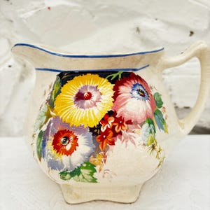 May include: Cream ceramic jug with a blue rim and handle. The jug is decorated with colourful floral designs in yellow, red, pink, and blue. The jug has a small spout and sits on a small base.
