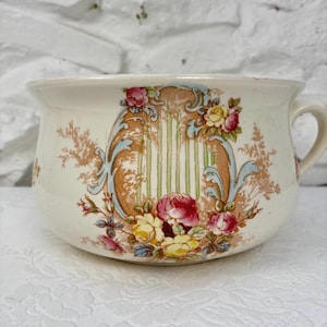 May include: Vintage cream-coloured chamber pot with a floral design. The pot has a central decorative panel with vertical stripes, framed by blue and brown scrollwork, and decorated with pink and yellow roses.