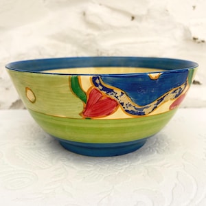 May include: A ceramic bowl featuring a hand-painted design with a wide blue rim and base. The exterior displays horizontal bands of light green and yellow, accented by abstract shapes in deep blue, bright red, and small yellow circles.