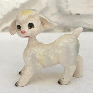 May include: A small ceramic figurine of a lamb with a glossy off-white finish. The lamb features large dark eyes, a small pink nose, and subtle yellow accents on the top of its head. It has four legs, a short tail, and dark-tipped hooves.