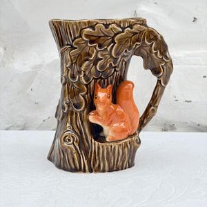 May include: A ceramic pitcher shaped like a brown tree trunk with textured oak leaves and a glossy finish. A small, orange squirrel is sculpted in relief on the front of the trunk, and the handle is formed by a branch extending from the side.