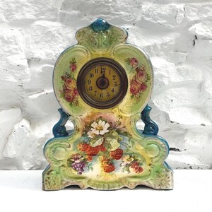 May include: A ceramic mantel clock with a pale green and yellow glazed finish, featuring a central circular clock face with black numerals. The body is decorated with painted floral motifs, including pink roses, white blossoms, red berries, and purple grapes.