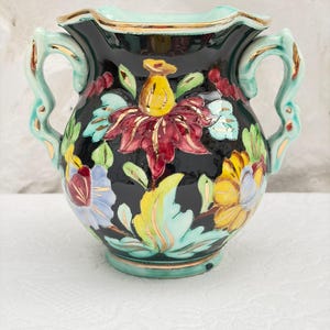 Könnte beinhalten: Ein schwarzer Keramikkrug mit zwei Henkeln und einem handgemalten Blumenmuster in Rot-, Gelb-, Blau- und Hellgrüntönen. Der Rand und die Henkelkanten sind mit Gold verziert, und der Körper weist eine glänzende, reflektierende Oberfläche auf.