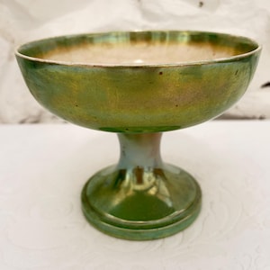 May include: A pedestal bowl featuring a lustrous, iridescent green finish with subtle golden undertones. The bowl has a wide, shallow basin supported by a sturdy, flared circular base, creating a smooth, reflective surface throughout the exterior.