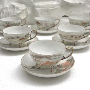 Op de afbeelding: Een set van zeven witte porseleinen kopjes en schotels. Elk kopje en schotel heeft een landschapsontwerp met gedempte tinten oranje, bruin en groen. De kopjes hebben goudkleurige handvatten en randen, en de schotels hebben een bijpassende gouden rand.