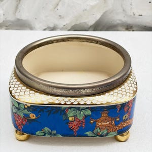 Può includere: Una ciotola ovale in porcellana con bordo metallico e quattro piccoli piedini dorati. L'esterno presenta un vivace sfondo blu decorato con un motivo colorato di bacche rosse, foglie verdi e una struttura a pagoda con squame bianche.