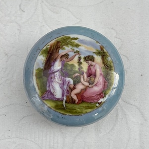 Könnte beinhalten: Eine runde Porzellan-Schmuckdose mit blau-weißem Design. Der Deckel zeigt eine bemalte Szene mit drei Figuren in einer Gartenlandschaft. Die Figuren sind eine Frau in einem lila Kleid, eine Frau in einem rosa Kleid und ein Kind in einem rot-weißen Outfit.