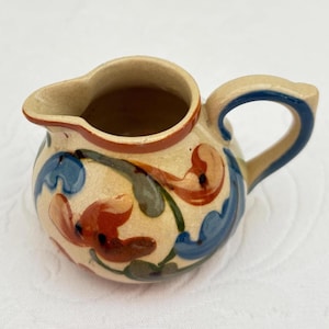May include: A small, ceramic creamer with a beige base, adorned with red, blue, and green floral patterns. The handle and rim are accented with blue and red, adding a touch of color to this decorative piece.