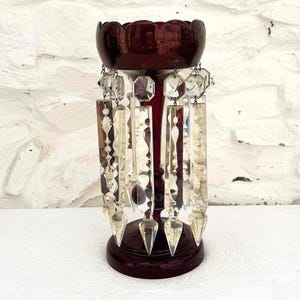Puede incluir: Un portavelas de cristal rojo oscuro con un borde superior festoneado y una base circular. La pieza está adornada con múltiples cristales de vidrio transparentes y facetados que cuelgan del borde, cada uno con una punta y un centro decorativo.