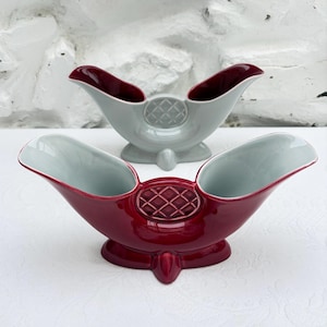 May include: Two ceramic boat-shaped vessels with dual openings and a central circular medallion featuring a raised diamond grid pattern. One vessel is deep burgundy red with a light interior, and the other is light grey with a dark interior.