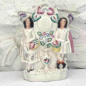 Puede incluir: Una figura de cerámica vintage con dos figuras con vestidos y sombreros blancos, sosteniendo cestas de flores. La figura tiene un diseño floral central y está pintada con acentos rosas, azules y verdes. La base es blanquecina.