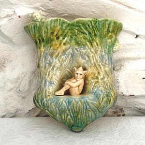 May include: A ceramic wall pocket planter featuring a sculpted tree trunk design with green and blue glaze. A small, light-colored, seated figure with pointed ears is molded into the center, with another small figure peering over the top edge.