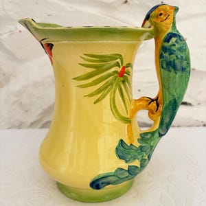 Może przedstawiać: Żółty ceramiczny dzbanek z ręcznie malowanym wzorem zielonych liści palmowych i trójwymiarowym uchwytem w kształcie papugi. Papuga jest pomalowana w odcieniach zieleni, żółci i błękitu, a jej pióra ogona rozciągają się wzdłuż boku dzbanka.