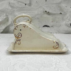 Puede incluir: Mantequera de cerámica color crema con tapa y asa. La base es rectangular y la parte superior curvada. Adornos dorados en forma de volutas decoran los lados y la parte superior, añadiendo un toque de elegancia a esta vajilla.