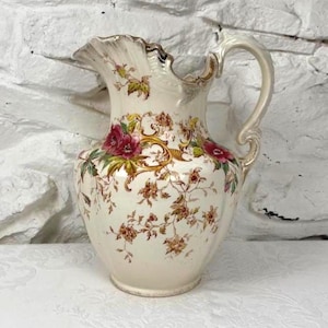 Może przedstawiać: Kremowy ceramiczny dzban z falistym brzegiem i ozdobnym uchwytem. Dzban jest zdobiony bordowymi i różowymi wzorami kwiatowymi, zaakcentowanymi złotymi detalami. Klasyczny element do serwowania napojów lub jako przedmiot dekoracyjny.