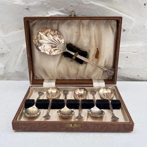 May include: A set of seven silver-toned spoons with shell-shaped bowls, presented in a brown rectangular case with a cream-coloured satin lining. One large serving spoon is secured in the lid, while six smaller spoons are arranged in the base.
