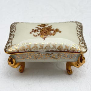 Puede incluir: Un joyero rectangular de porcelana con base y tapa de color crema. La tapa presenta un ramo de flores dorado central y bordes de filigrana dorada. Los lados están decorados con enredaderas doradas y se apoya sobre cuatro patas doradas.