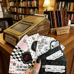 Puede incluir: Una mano sostiene un abanico de tarjetas decorativas con varios dise&ntilde;os, incluyendo patrones florales, una calavera y el texto "I don't watch porn. I read it like a lady, thankyou." Un terminal de pago dorado y de madera se encuentra en el fondo.