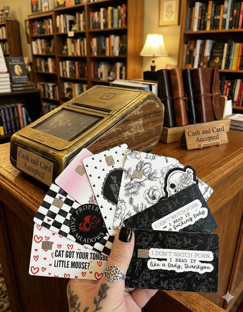 Puede incluir: Una mano sostiene un abanico de tarjetas decorativas con varios dise&ntilde;os, incluyendo patrones florales, una calavera y el texto "I don't watch porn. I read it like a lady, thankyou." Un terminal de pago dorado y de madera se encuentra en el fondo.