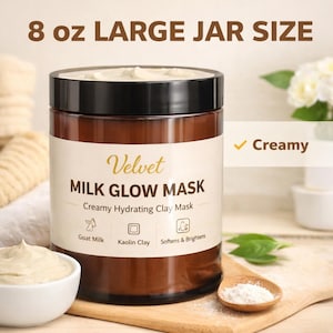 May include: A brown jar of Velvet Milk Glow Mask, 8 oz size, with a black lid. The label reads "Creamy Hydrating Clay Mask" and lists goat milk and kaolin clay as ingredients. A small bowl of the mask and a wooden spoon with powder are also visible.