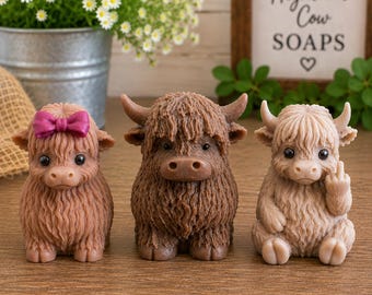 May include: Three sculpted Highland cow figures made of soap. The left figure is light brown with a pink bow. The center figure is dark brown with horns. The right figure is beige with horns, holding up its middle finger. Each has dark eyes.
