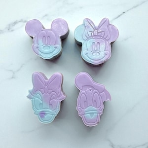 May include: Four handmade soaps in the shape of Disney characters: Mickey Mouse, Minnie Mouse, Daisy Duck, and Donald Duck. Each soap is lavender and blue, with a marble-like effect. The soaps are arranged on a white surface.