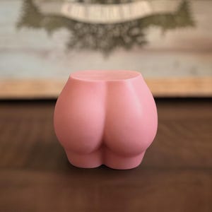 May include: A pink, butt-shaped decorative object. The item is a solid color and has a smooth surface. The object is sitting on a brown surface.