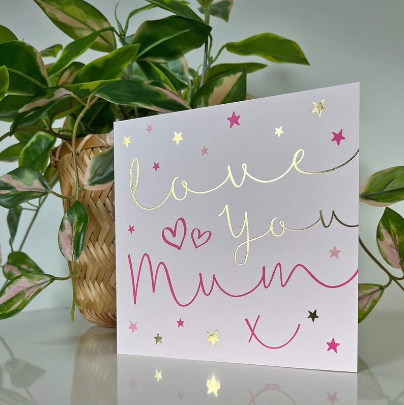 May include: A pink and gold card with the text "love you mum" and a pink heart. The card is decorated with gold stars.