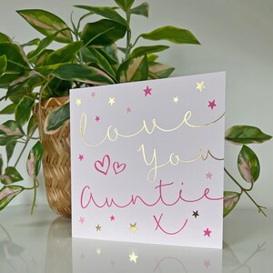 May include: A white card with pink and gold lettering that says "love you auntie x" with pink hearts and gold stars scattered around the text.