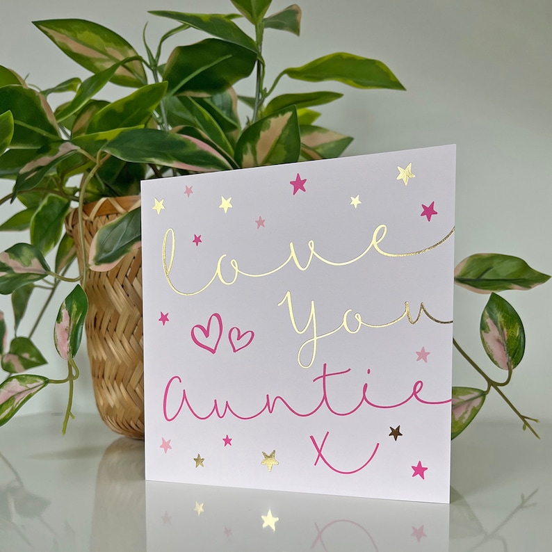 May include: A white card with pink and gold lettering that says "love you auntie x" with pink hearts and gold stars scattered around the text.