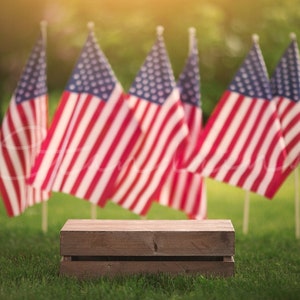 May include: A wooden crate sits in front of a blurred background of American flags waving in the wind. The flags are red, white, and blue.