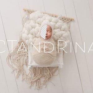 May include: A newborn baby swaddled in a cream-coloured blanket, lying on a white woven cushion with a white background. The baby is wearing a white knitted hat and has a neutral expression. The text "CTANDRINA" is visible in the image.